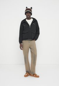 Άτομο με ριγέ πλεκτό καπέλο με αυτιά, μαύρο μπουφάν με φερμουάρ, λευκό πουκάμισο, μπεζ τζιν και καφέ αθλητικά παπούτσια, στέκεται μπροστά σε λευκό τοίχο.