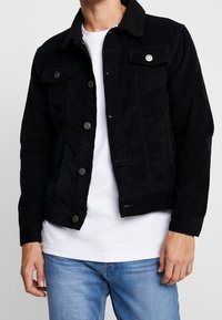 Man med svart manchesterjacka med knappar, vit t-shirt under och ljusblå jeans, som står framför en neutral bakgrund.