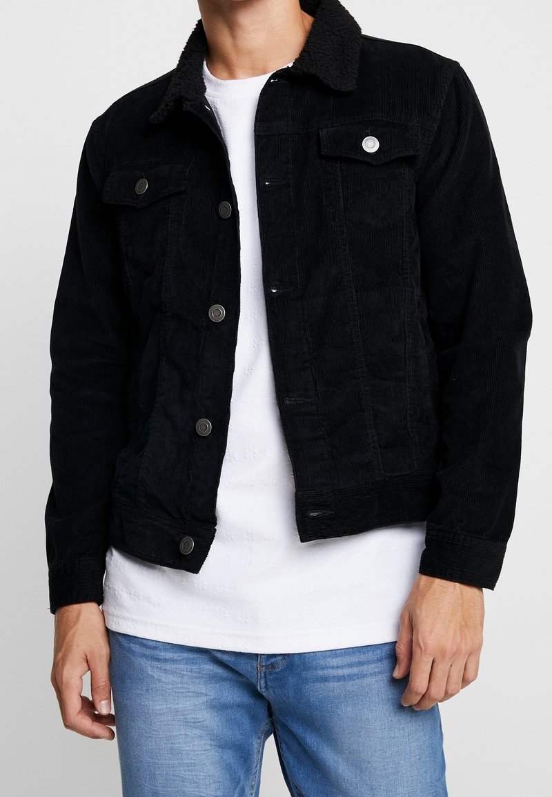 Man med svart manchesterjacka med knappar, vit t-shirt under och ljusblå jeans, som står framför en neutral bakgrund.