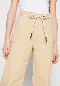 Béžové bavlněné kalhoty s širokým střihem, s bílým stahovacím pasem s černými akcenty a bočními kapsami. Hladká textura, minimalistický design.