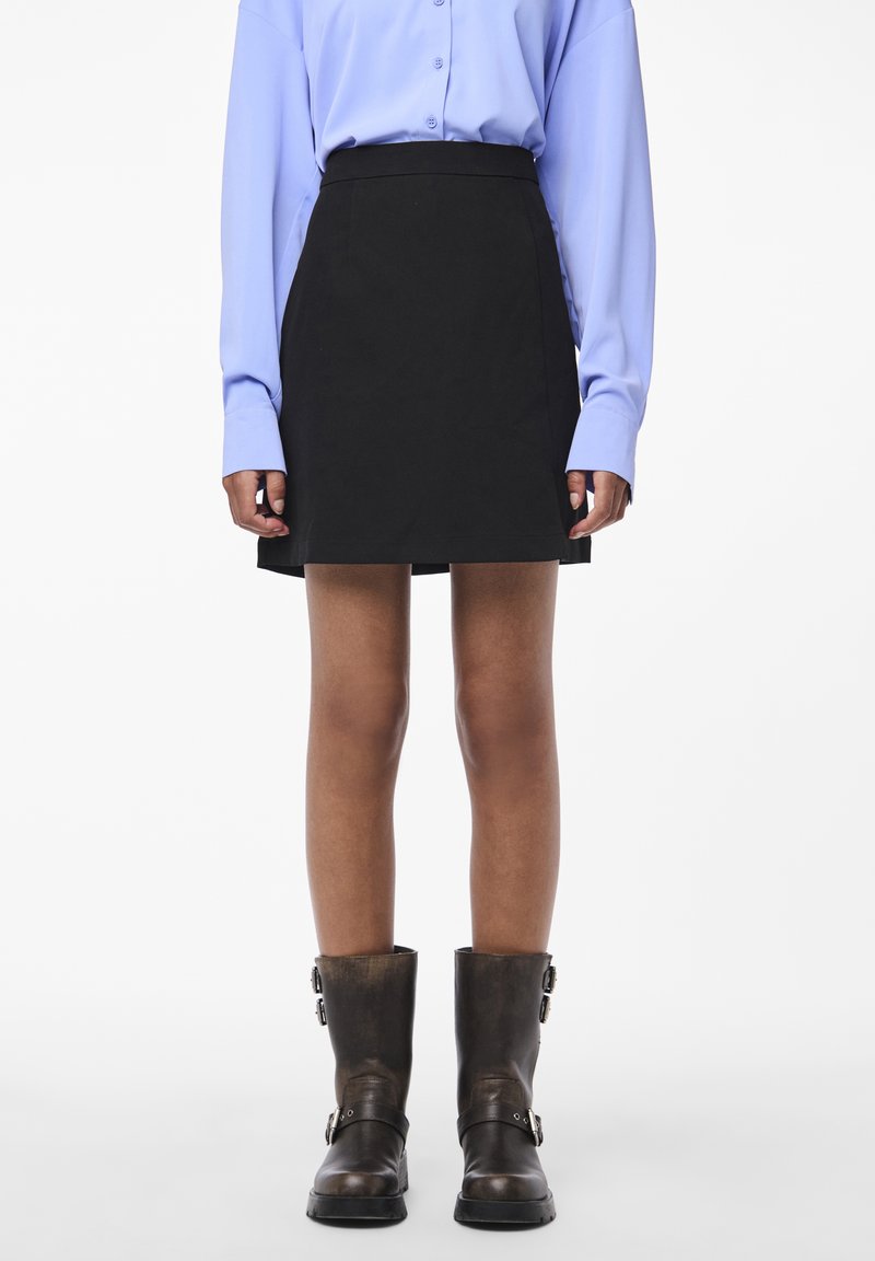 Musta mini-hame, jossa on istuva vyötärö ja suora siluetti. Yhdistetty vaalean sinisen napitettu paita ja ruskeat nilkkurit.