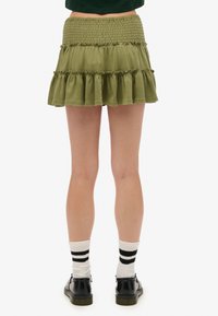 Olijfgroene, gelaagde mini-jurk met smocktail, gerafelde randen en zachte stof. Gecombineerd met zwarte schoenen en knielange gestreepte sokken.