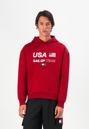 Rød hettegenser med hvit tekst "USA SAIL GP TEAM," med et flagg-design. Framlomme og ribbestrikkede mansjetter. Modellen har på seg svarte shorts.
