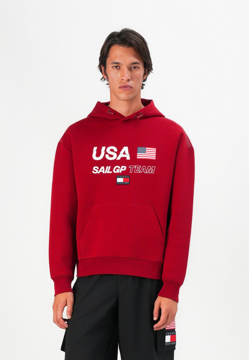 Rød hættetrøje med hvid tekst "USA SAIL GP TEAM," med et flaggrafik. Forlomme og ribbede manchetter. Modellen har sorte shorts på.