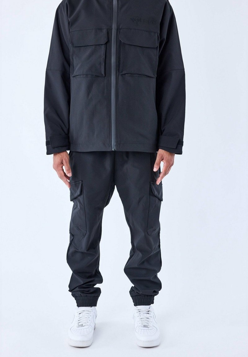 Veste zippée noire avec deux poches poitrine, pantalon cargo assorti avec poches à rabat, et baskets blanches sur une personne debout devant un fond uni.