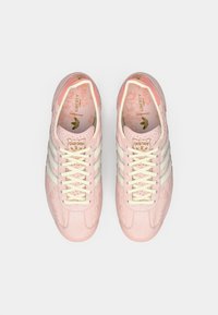 Világos rózsaszín sportcipők texturált felsőrésszel, fehér fűzővel és három kontrasztos csíkkal. A belső rész virágmintával és logórészletekkel díszített.