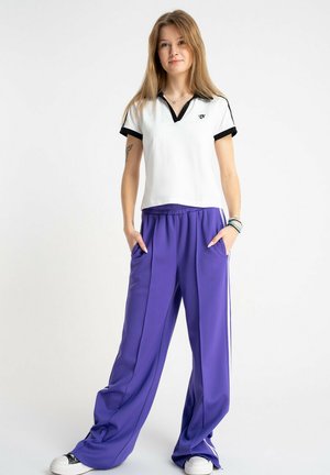 Młoda kobieta stojąca z rękami w kieszeniach, ubrana w białą koszulkę polo i luźne fioletowe spodnie dresowe z białymi paskami.