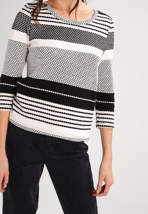 Femme portant un haut à manches longues à motif noir et blanc avec des rayures horizontales et des designs à points hexagonaux, associé à un pantalon noir.