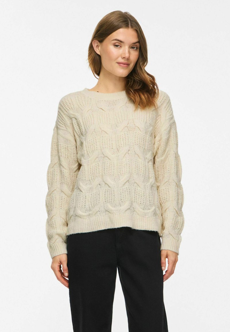 Femme portant un pull torsadé couleur crème et un pantalon noir, debout devant un fond blanc uni.