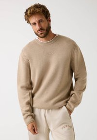 Sweatshirt beige con colletto a costine e logo ricamato "GUESS". Tessuto morbido, vestibilità rilassata, abbinato a pantaloni della tuta beige.