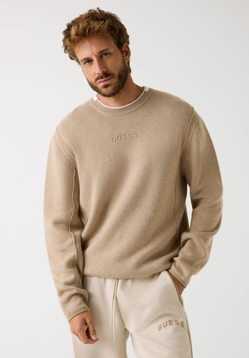 Sweatshirt beige con colletto a costine e logo ricamato "GUESS". Tessuto morbido, vestibilità rilassata, abbinato a pantaloni della tuta beige.