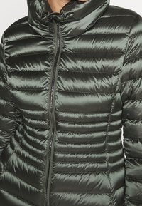 Mellanlång pufferjacka i mörkgrön nylon, med horisontell kviltad söm, hög krage och central dragkedjestängning med metall-accenter.