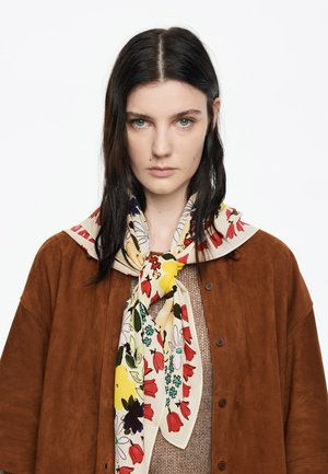 Pañuelo de seda con estampado floral que presenta un fondo beige con tulipanes rojos, acentos amarillos y hojas verdes, atado alrededor del cuello sobre una chaqueta marrón.