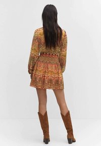 Vestido de manga larga, con estampado en naranja, amarillo y verde, con cintura ajustada y falda acampanada. Combinado con botas de ante marrón hasta la rodilla.
