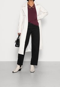 Długi biały płaszcz na bordowej bluzce z koronkowymi detalami, w połączeniu z czarnymi spodniami o szerokich nogawkach oraz kremowymi botkami za kostki z czarnymi czubkami.