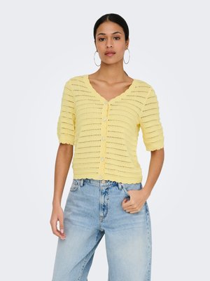 Donna che indossa un cardigan in maglia giallo a maniche corte con bottoni e bordi smerlati e jeans azzurri a vita alta, in piedi con la mano in tasca.