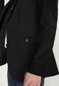 Schwarzer Blazer mit strukturiertem Design, standardmäßigen Revers, vier Knöpfen und einer Tasche mit einem schwarzen Logo-Detail an den Ärmeln. Glatte Stoffqualität.