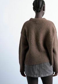 Personne aux cheveux tressés en chignon portant des boucles d'oreilles créoles argentées, un pull marron en matière duveteuse et une mini-jupe en denim gris, vue de dos.