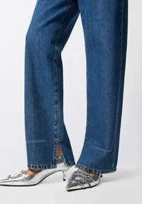 Srebrne cipele na petu uparene s plavim trapericama. Traperice imaju mali prorez na rubu, a cipele su sa špicastim vrhom i zlatnim detaljima.