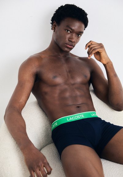 Черни боксерки на Lacoste с еластичен колан в зелено, седят на текстурирано кремаво кресло. Гладък тон на кожата, мускулесто тяло.