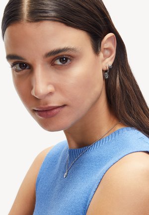 Silberne Ohrringe mit einem runden Anhänger, getragen zu einem blauen, gerippten, ärmellosen Top. Glatte Textur und unauffälliges Design. Nahansicht im Porträtformat.
