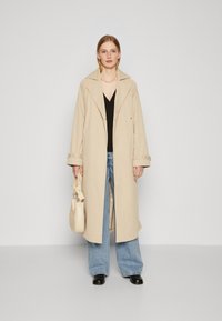 Beige trenchcoat med bred krage, knappåpning og justerbare ermer. Bäres over en svart topp og lyseblå jeans, kombinert med en kremfarget veske.