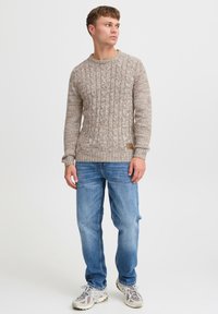 Beige kabelstrikket genser med rund hals, kombinert med lyseblå jeans og mønstrede grå sneakers. Merkbar sømdetalj og logo.