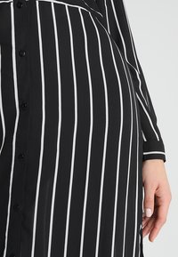 Robe noire à rayures blanches verticales, boutons sur le devant, manches longues et une main avec un vernis à ongles clair reposant sur le côté.
