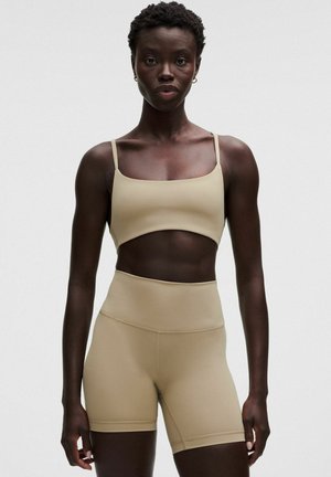 Atletisk kvinde med kort hår iført beige sports-bh og cykelshorts med høj talje, står og vender fremad mod en ensfarvet baggrund.