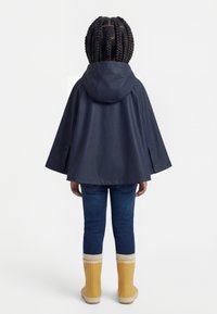 Dijete u tamnoplavom kišnom ogrtaču s kapuljačom, tamnim trapericama i žutim čizmama za kišu, stoji okrenuto leđima na bijeloj pozadini.
