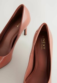 Zapatos de tacón alto de charol rosa con una forma elegante, puntera afilada y un tacón de aguja de 10 cm. El interior presenta un logotipo dorado.