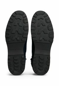 Une paire de bottes noires à semelle en caoutchouc avec des motifs de traction profonds et le logo "Tommy Hilfiger" sur la zone du talon.