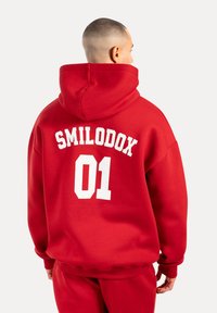 Sudadera roja de gran tamaño hecha de algodón, con el texto blanco "SMILODOX 01" en la parte posterior. Incluye una capucha central y puños acanalados.