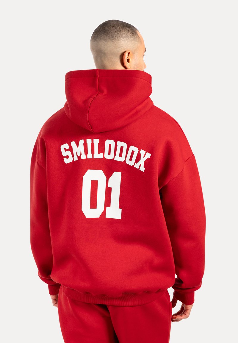 Sudadera roja de gran tamaño hecha de algodón, con el texto blanco "SMILODOX 01" en la parte posterior. Incluye una capucha central y puños acanalados.