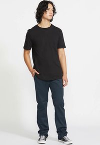 T-shirt preta com gola redonda, mangas curtas, combinada com calças azul escuro. O modelo usa ténis pretos e brancos. Design simples e ajustado.