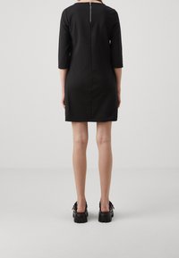 Robe noire à manches courtes avec une fermeture éclair apparente dans le dos. Tissu lisse, silhouette ajustée et ourlet droit. Associée à des chaussures noires épaisses.