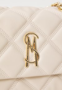 Tikkimusteriga beež nahast käekott kuldsete detailidega, millel on omavahel seotud monogramm-logo ja nähtavad õmblusdetailid.
