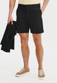 Czarne krótki spodenki wykonane z lekkiego materiału z zapięciem na guzik, w połączeniu z beżową koszulą i brązowymi trampkami. Prosty, czysty design.