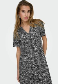 Schwarzes Kleid mit weißem Blattmuster, kurzen Ärmeln, V-Ausschnitt und gerafftem Bund. Leichtes Material mit weicher Textur.