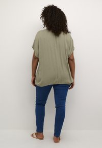 Mujer con cabello rizado que lleva una blusa ligera de oliva suelta, jeans azules y sandalias beiges, de pie frente a una pared blanca lisa.