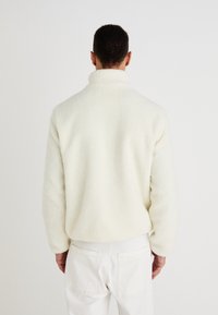 Casaco de lã branca com gola alta, tecido texturizado e um corte descontraído. Usado sobre calças brancas, apresentando um design minimalista.