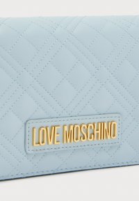 Φωτεινό γαλάζιο καπιτονέ πορτοφόλι με χρυσή πλακέτα "LOVE MOSCHINO", ομαλή υφή και ραφές σε σχέδιο διαμαντιού.