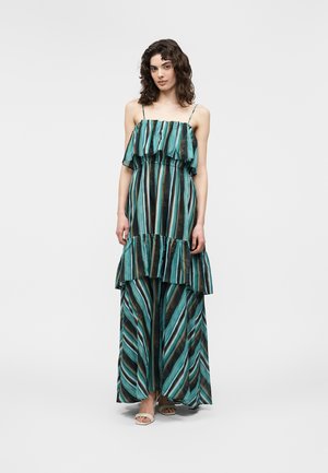 Vrouw draagt een mouwloze maxi-jurk met strepen in teal, zwart en bruin, met laagjes en dunne bandjes, staand tegen een effen achtergrond.