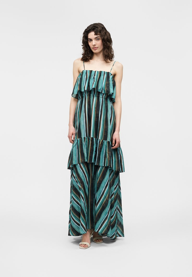 Γυναίκα που φοράει ένα αμάνικο μακρύ φόρεμα σε τυρκουάζ, μαύρο και καφέ ρίγες με στρώσεις βολάν και λεπτές τιράντες, στέκεται μπροστά σε ένα απλό φόντο.