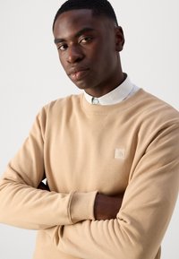Sudadera beige con cuello redondo y puños acanalados. Presenta un sutil logo texturizado en el lado izquierdo del pecho. Fabricada en suave algodón y con un ajuste relajado.