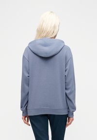 Άτομο με μακριά ξανθά μαλλιά, που φοράει ανοιχτό μπλε φούτερ με κουκούλα και σκούρο τζιν, γυρισμένο πλάτη σε ένα απλό λευκό φόντο.