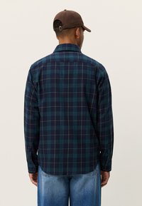 Καρό πουκάμισο σε ναυτικό μπλε και πράσινο, κατασκευασμένο από μαλακό ύφασμα, με γιακά που κουμπώνει, μακριά μανίκια και μανσέτες με κουμπιά. Φοριέται με ανοιχτομπλε τζιν.