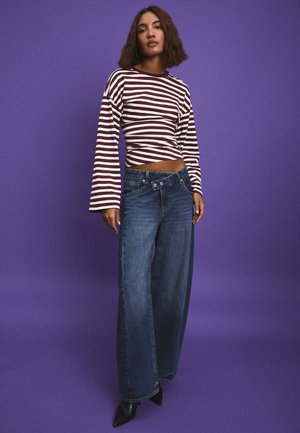Femme portant un chemisier à manches longues rayé bordeaux et blanc et un jean bleu taille haute à jambes larges, debout devant un fond violet.