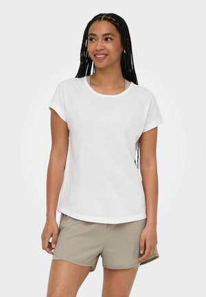 Woman with long braids wearing a white short-sleeve shirt and beige shorts, smiling and standing against a plain background.
