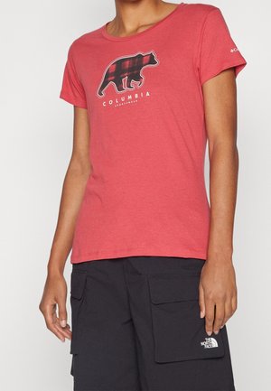 Persoon draagt een rood Columbia t-shirt met een geruit berenlogo en zwarte North Face cargoshorts met grote voorzakken.